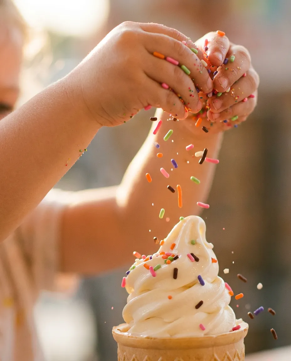A child's hands sprinkling colorful toppings onto a swirl of soft ice cream.